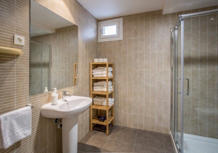 Baño moderno con azulejos beige, lavabo, espejo, ducha y toallero de madera.