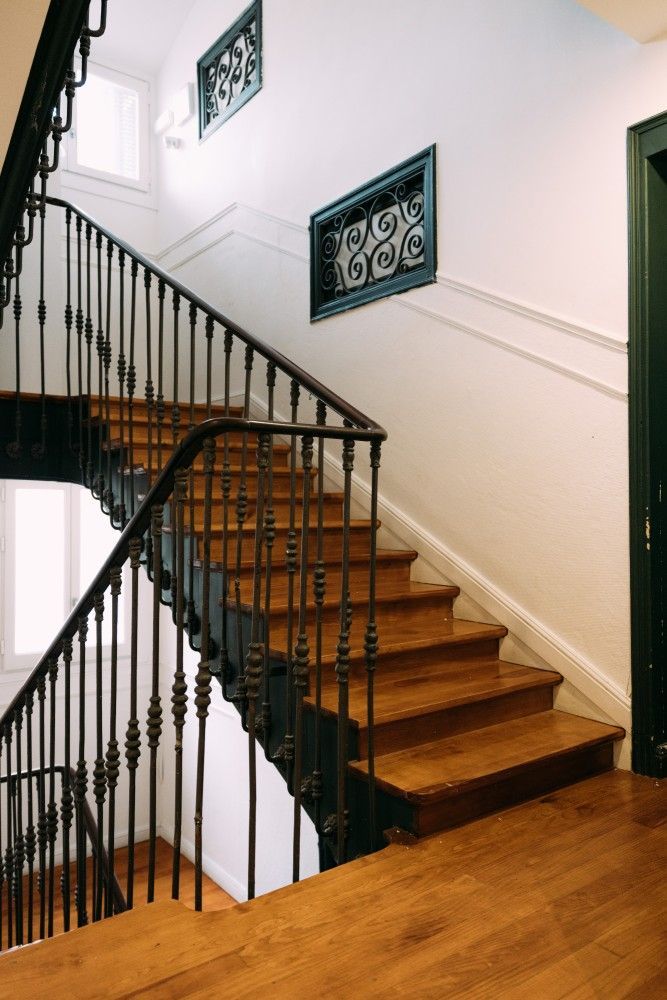 Escalera de madera con barandilla de hierro forjado negro y cuadros enmarcados en un luminoso pasillo interior.