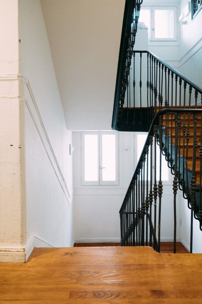 Escalera con barandilla de hierro negro, paredes blancas y suelo de madera cálida, iluminada por una pequeña ventana.