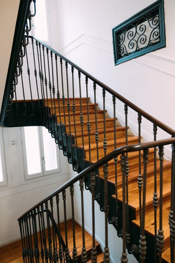 Escalera interior con peldaños de madera, barandillas negras y paredes blancas, junto a una ventana y una reja decorativa.