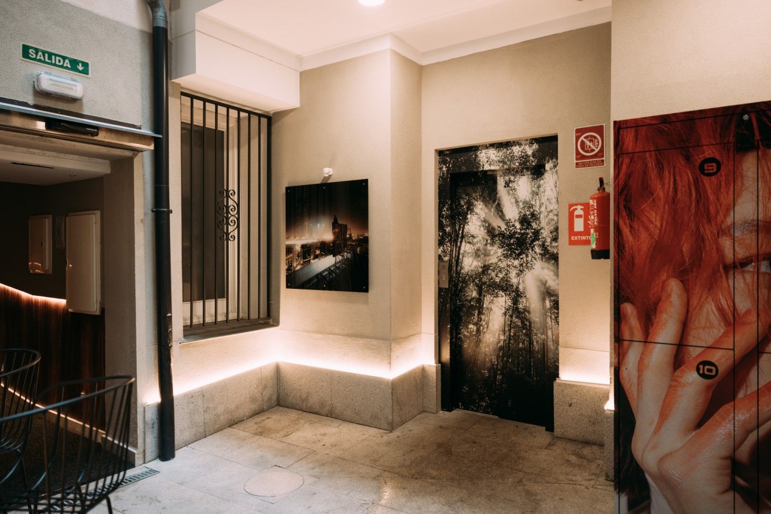 Pasillo con poca luz, puertas cubiertas de obras de arte, una ventana con barrotes y una luz cálida sobre paredes de color.