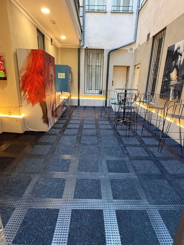 Estrecho pasillo exterior con alfombra azul, sillas, un tapiz rojo y una puerta azul bajo luces brillantes.