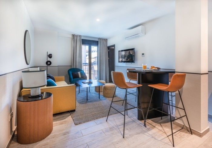Sala de estar de apartamento moderno con sillas azules y naranjas, una mesa de bar, TV y puertas que dan al balcón.