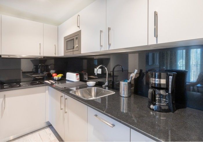 Cocina moderna con gabinetes blancos, encimeras negras, fregadero, cafetera y luz natural proveniente de la ventana.
