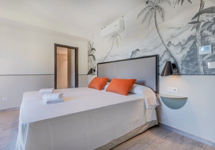 Dormitorio moderno con cama blanca, almohadas naranjas y papel tapiz mural de palmeras.