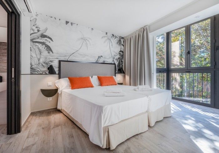 Dormitorio moderno con cama, almohadas naranjas, mural floral en la pared y luz natural que entra por grandes ventanales.