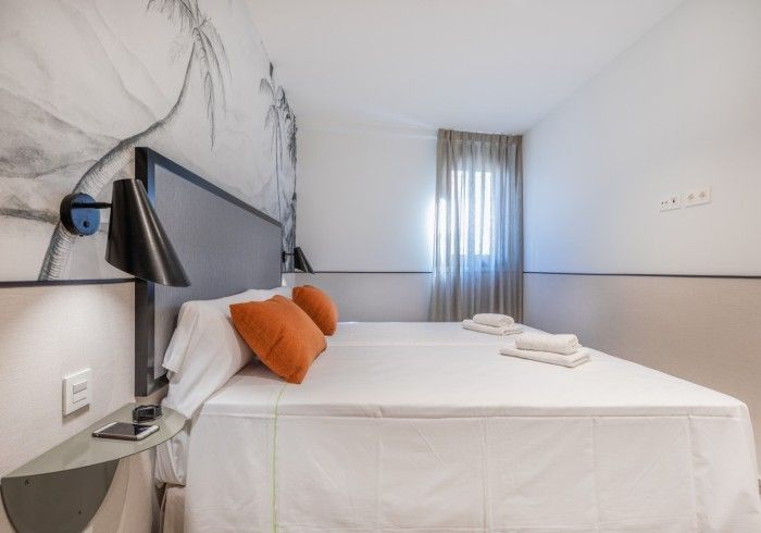 Habitación pequeña con cama doble, televisor montado en la pared, almohadas naranjas y ventana con cortinas transparentes.