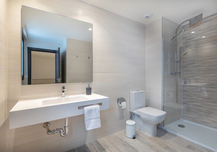 Baño moderno con lavabo suspendido, espejo grande, inodoro y ducha con mampara de cristal en tonos gris claro.