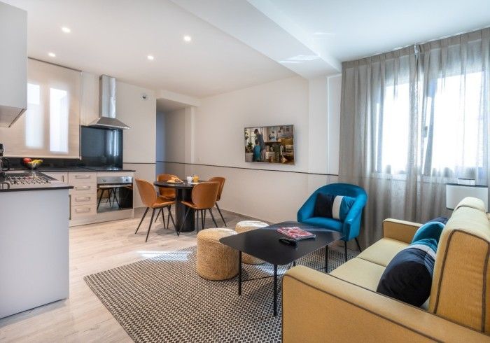 Salón moderno de planta abierta con cocina integrada, sofá beige, sillón azul y grandes cortinas en las ventanas.