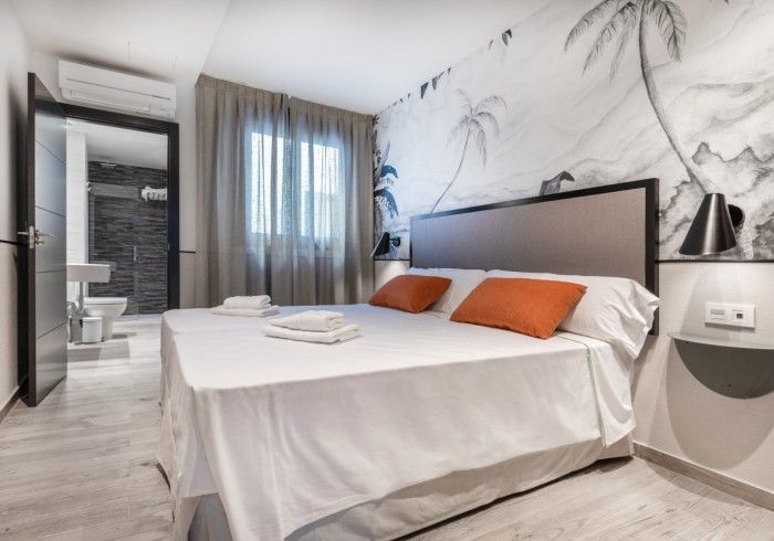Dormitorio moderno con cama blanca, almohadas naranjas, pared mural y puerta del baño visible