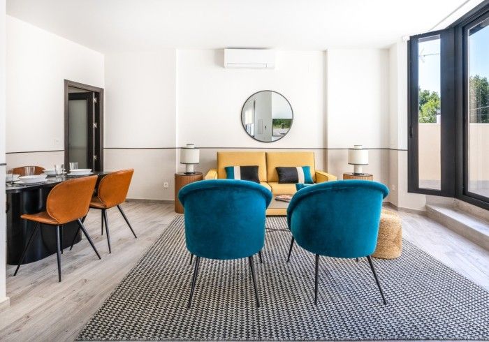 Luminoso salón moderno con sillones color turquesa, sofá amarillo, espejo redondo y grandes ventanales.