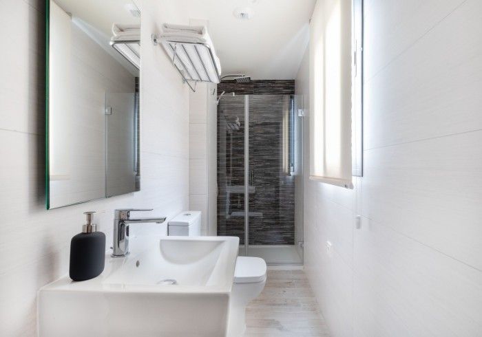 Baño moderno y luminoso con lavabo blanco, espejo, inodoro, ducha y luz de ventana.