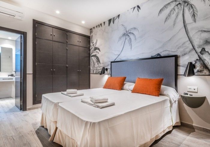 Dormitorio moderno con una cama grande, almohadas naranjas, armario oscuro y mural de palmeras en la pared.