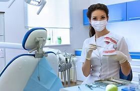 Dentist in mask and gloves, holding a teeth model, in a blue and white dental office.