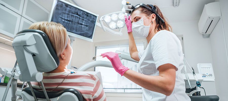 A dentist in mask and pink gloves examines a patient in a dental chair. A dental X-ray is visible on a monitor.