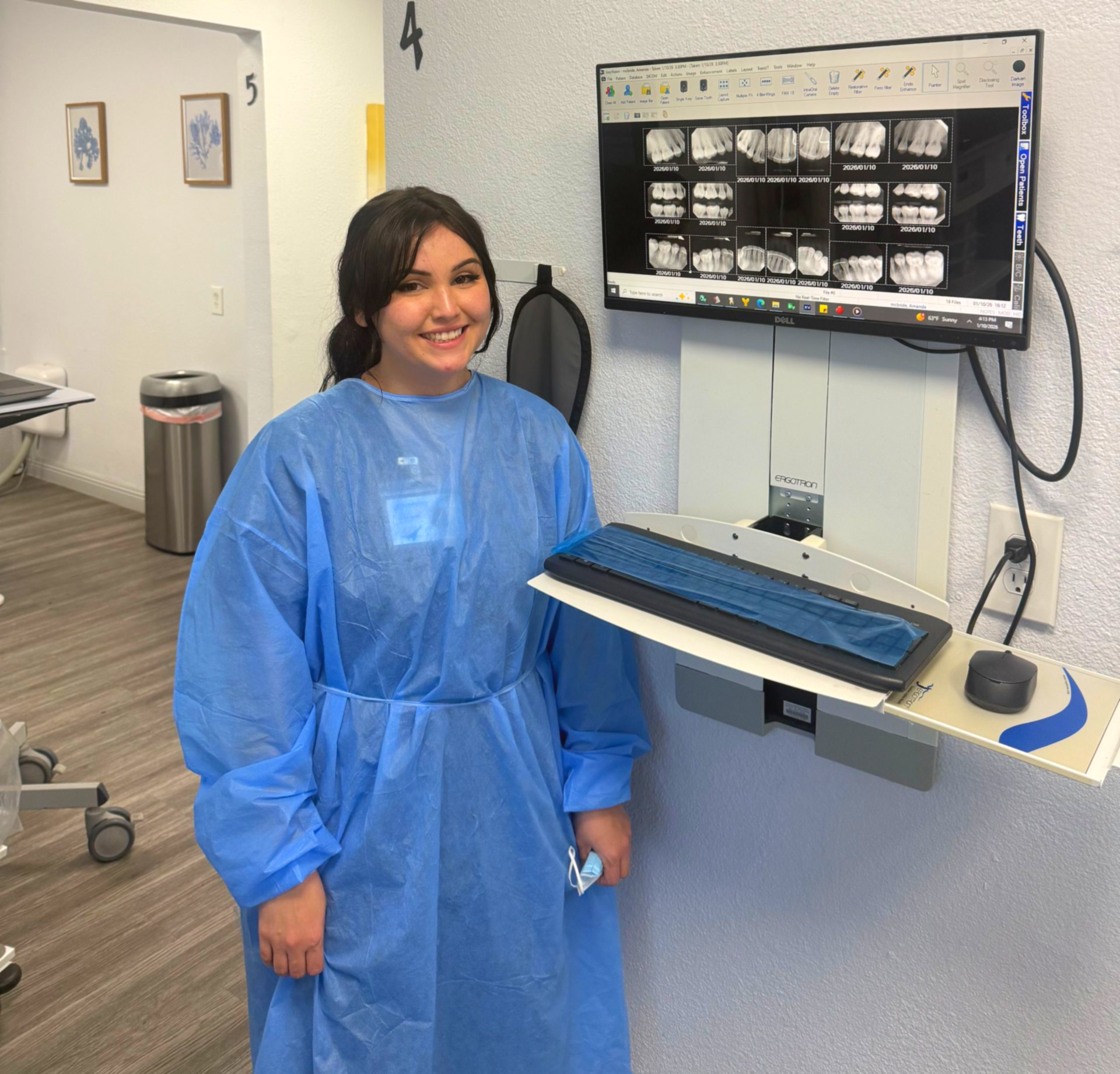 Dentist in blue scrubs examining a young patient's teeth in a dental office. The patient wears a lead apron.
