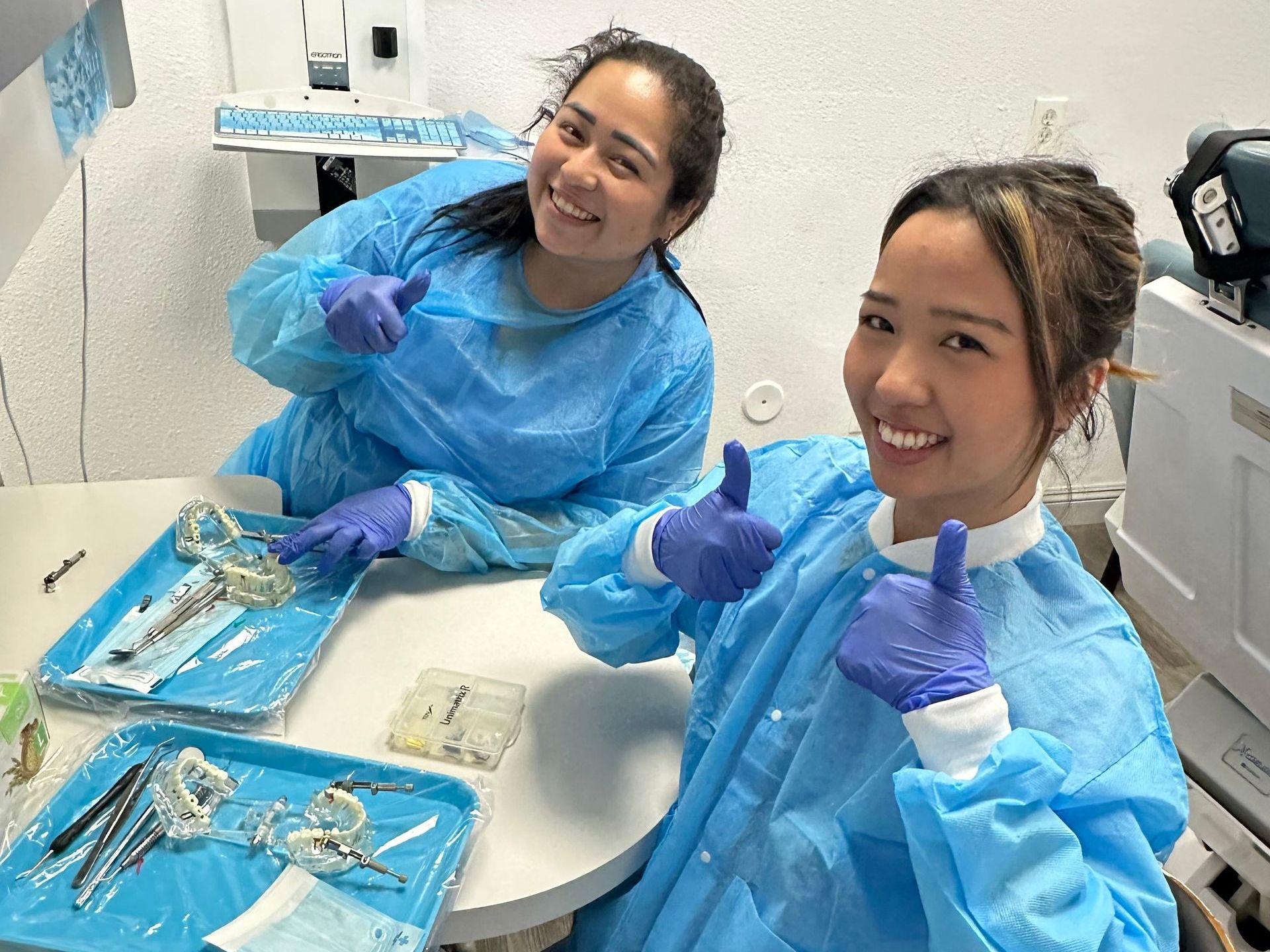 Two smiling women in blue scrubs giving thumbs-up in a dental office.