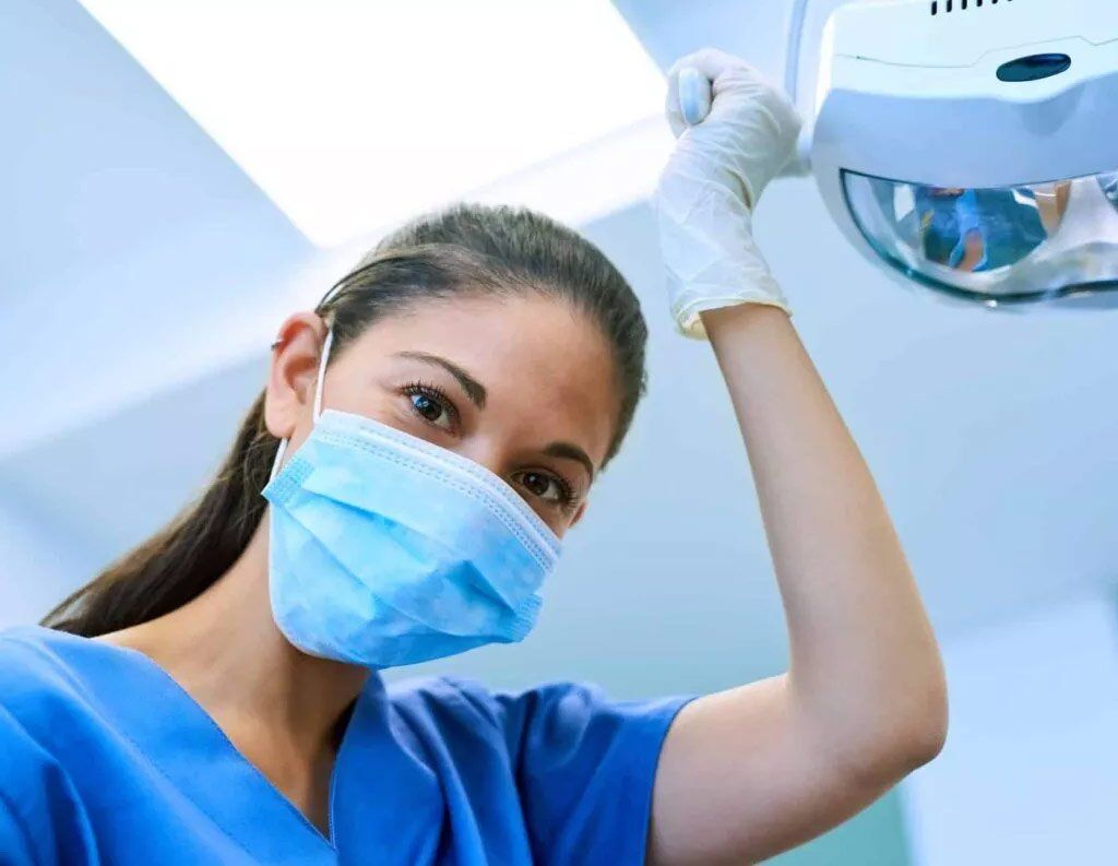 Dentist in blue scrubs and mask adjusting overhead light, looking down.