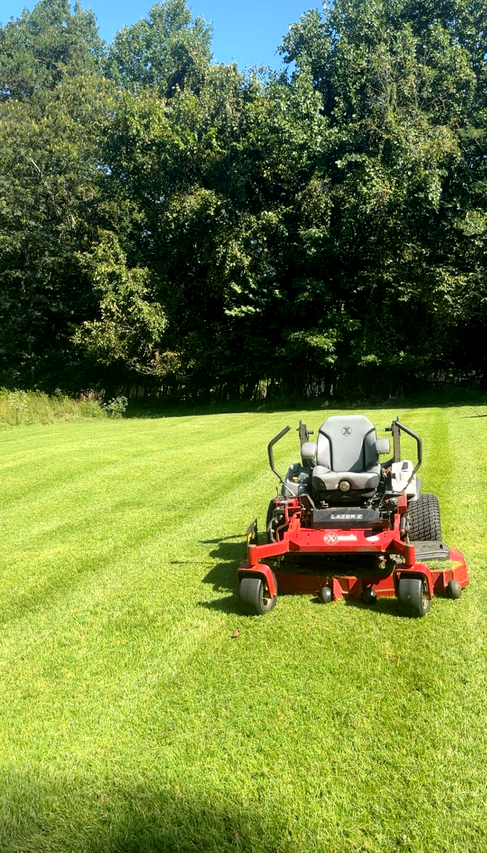 Red zero-turn mower mowing a green lawn with a backdrop of trees under a blue sky.