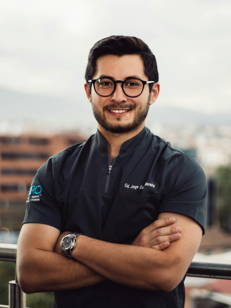 Man with glasses and arms crossed, smiling in medical scrubs, outdoors.
