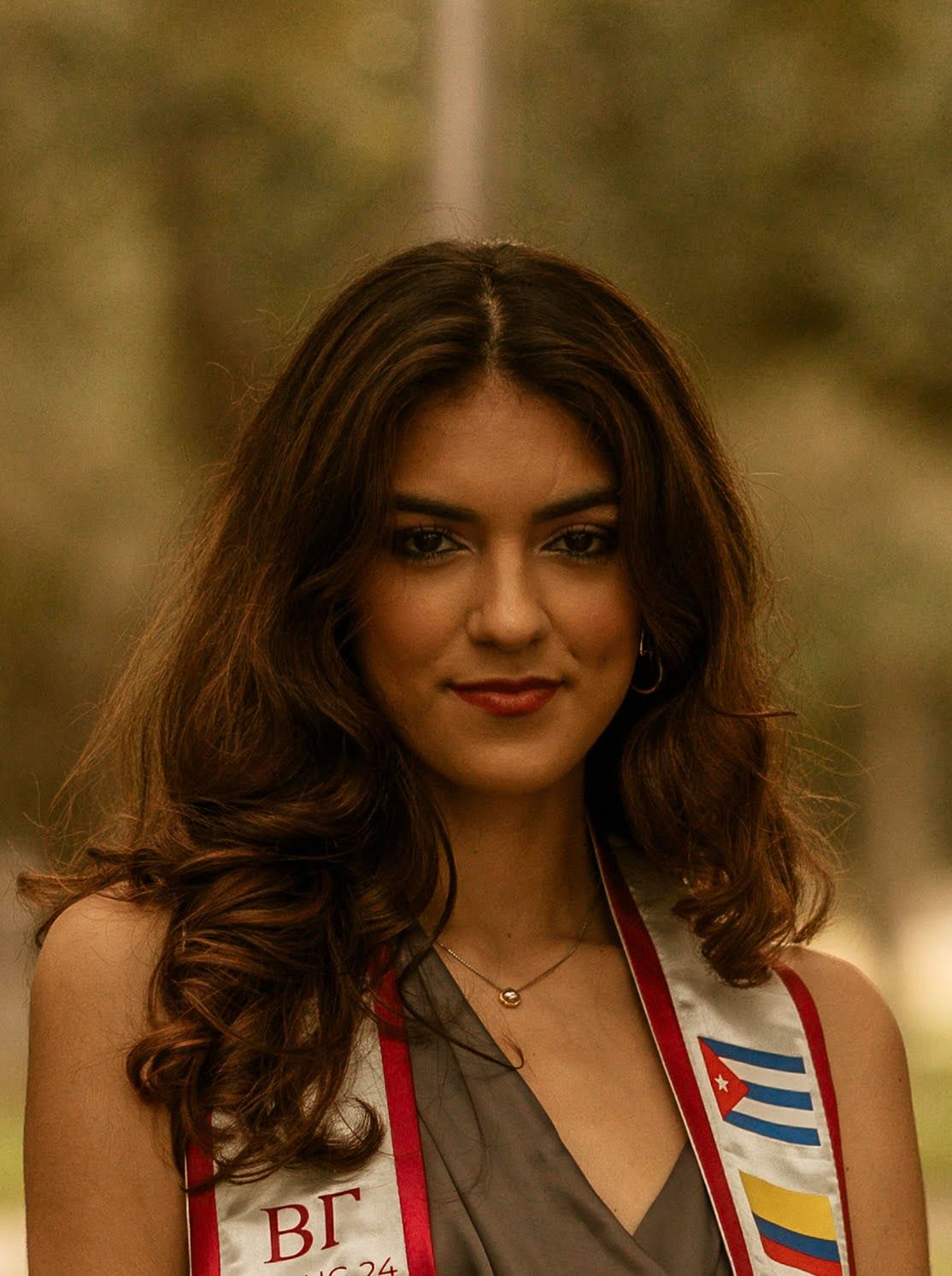 Woman with brown hair wearing a graduation sash, smiling at the camera, outdoors.