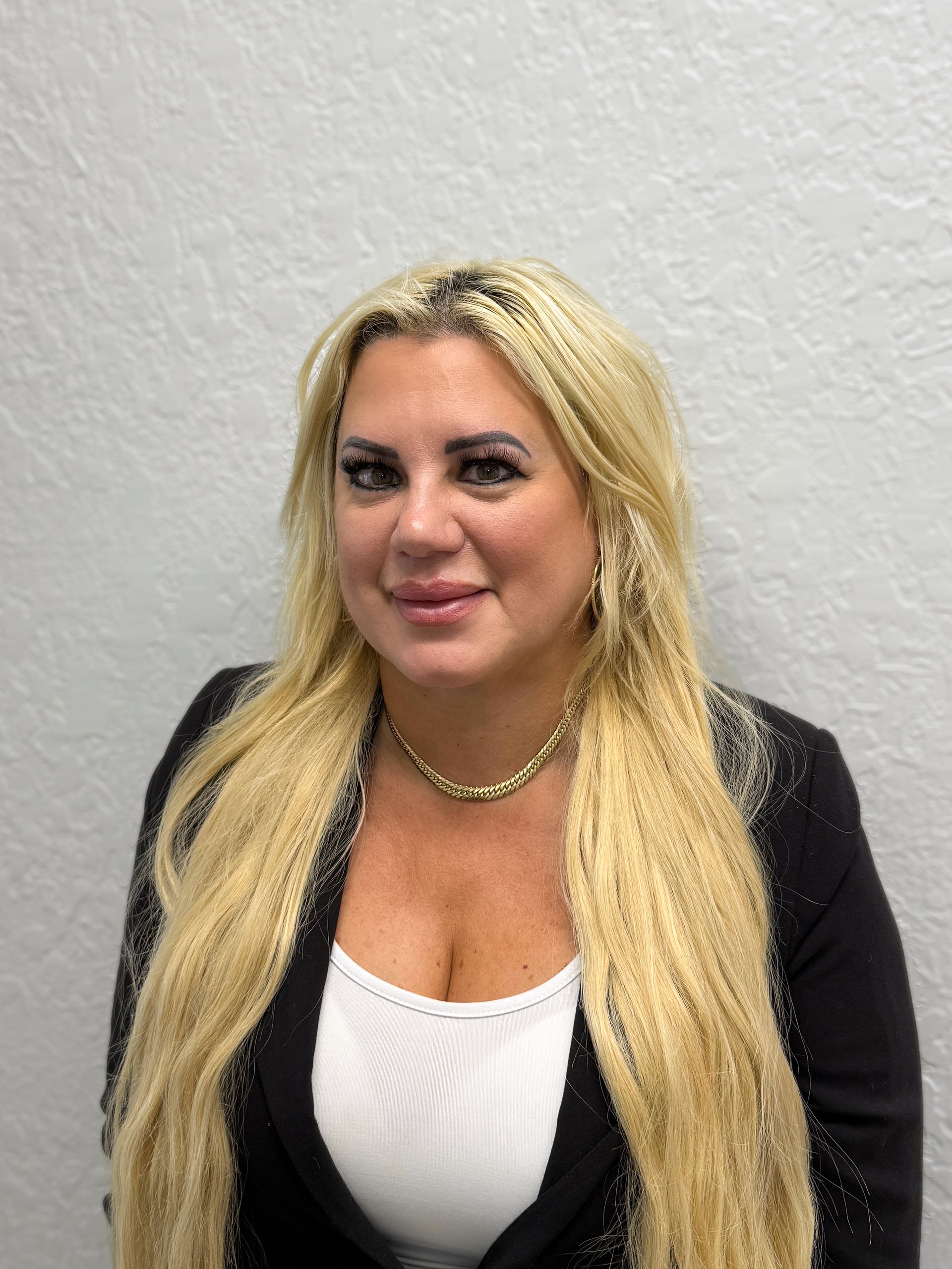 Blonde woman with pale skin, dark eyebrows, and a slight smile, wearing a gold necklace, against a black background.