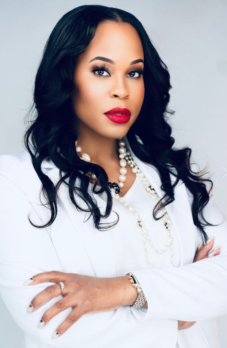 A woman wearing a white jacket and red lipstick is standing with her arms crossed.