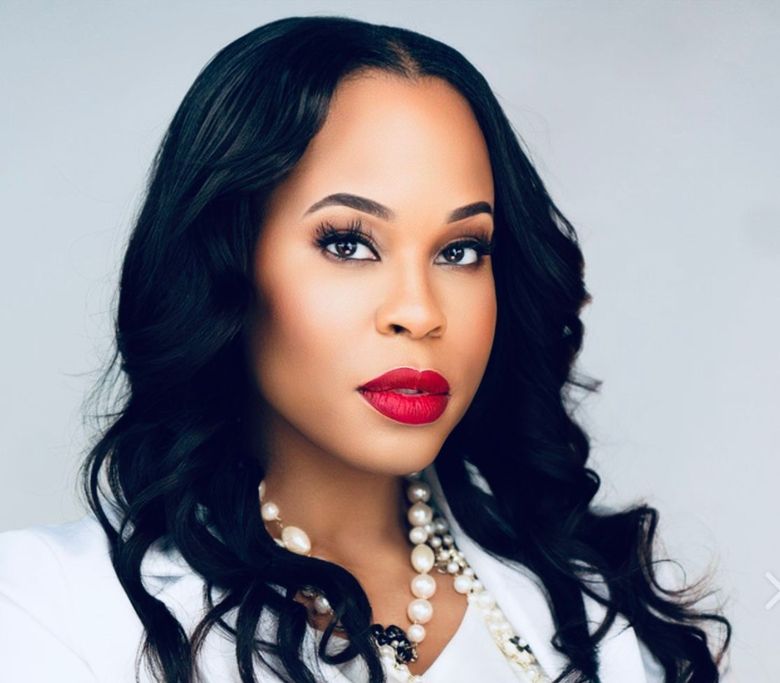 A woman wearing a white jacket and red lipstick is standing with her arms crossed.