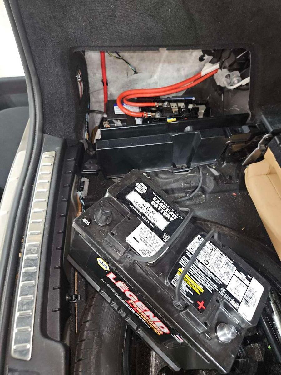 Car trunk interior with car battery on the floor, black container and wiring visible. | Accomplished Automotive