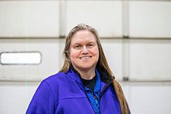 Woman with long brown hair smiling, wearing a blue jacket, in front of a white door.