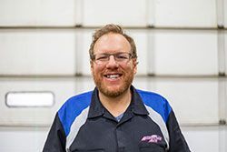 Man with glasses and beard, wearing a blue and black work shirt, smiling in front of a garage door.