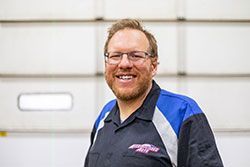 Man with glasses smiles, wearing a black shirt with blue and gray accents, in front of a white garage door.