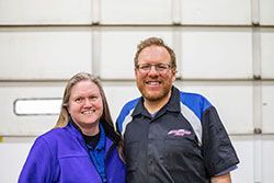 Woman and man smiling, posing in front of a white garage door. Both wear work shirts.
