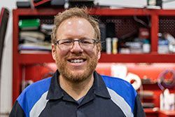 Man with glasses smiles in a garage, wearing a blue and black shirt.