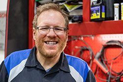 Man in blue and black shirt with glasses smiles in a workshop, red cabinet in background.