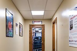 Hallway with closed door leading to an interior space; light walls, framed posters, and ceiling tiles.