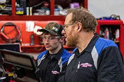 Two mechanics in a shop look at a computer. One wears a baseball cap and glasses. The other has glasses and a blue/black uniform.