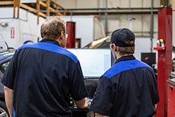 Two mechanics in blue and black uniforms examining a computer screen in a garage.