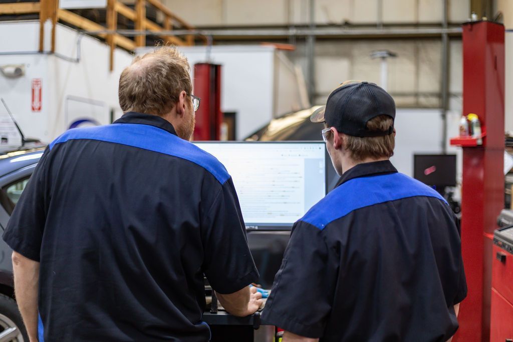 Two mechanics in blue and black uniforms examining a computer screen in a garage. | Accomplished Auto