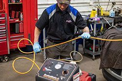 Mechanic in blue gloves, holding a yellow hose, working on a machine in a garage.