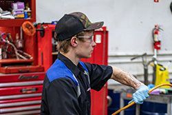 Mechanic in auto shop holding a yellow hose, wearing hat, safety glasses, and gloves.