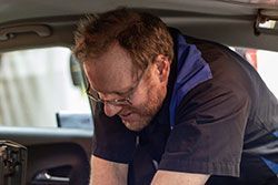 Mechanic working inside a car, wearing glasses and a blue and black uniform.