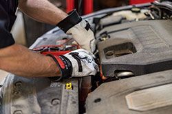 Mechanic wearing gloves working on a car engine, connecting jumper cables.