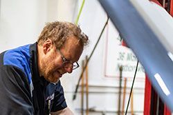 Mechanic with glasses working on car engine, wearing a blue and black uniform.