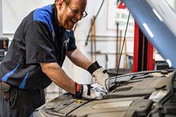 Mechanic in blue and black work shirt smiles while working on a car engine.