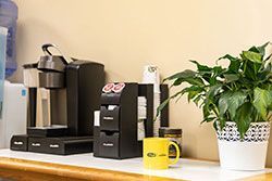 Coffee station with a Keurig, K-cup storage, cups, and a yellow mug, beside a potted green plant.