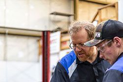 Two mechanics looking at something in a shop. One wears glasses and blue-accented shirt. The other has a cap and safety glasses.