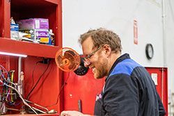 Mechanic in blue-shouldered work shirt smiles at a red workbench, wires and tools visible.