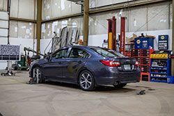 Dark blue car in a garage, undergoing alignment. Tools, shelves, and a lift are visible.