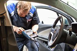 Mechanic in blue and black work shirt uses diagnostic tool inside a car.